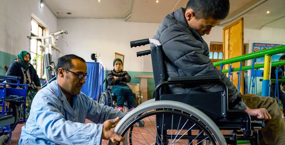 Personne en fauteuil roulant avec quelqu'un derrière qui semble réparer le fauteuil