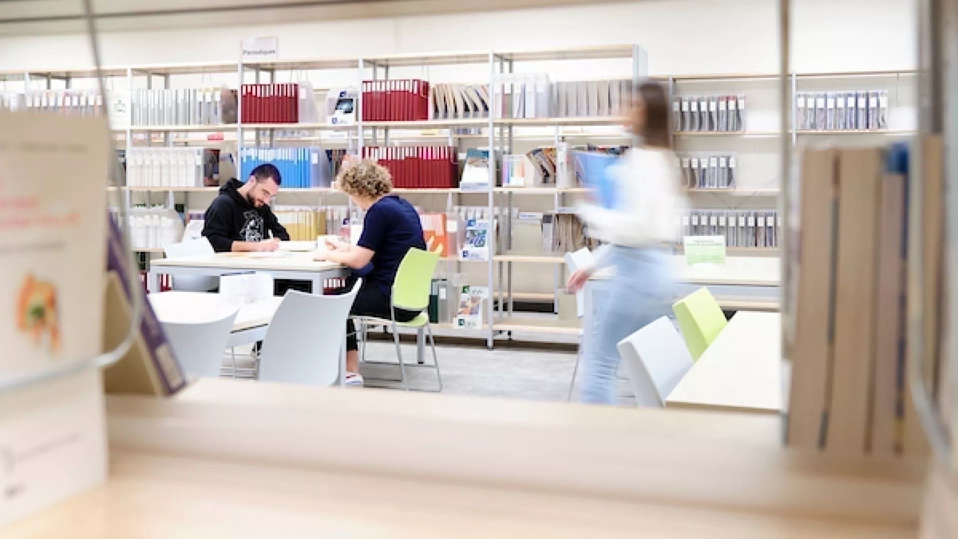 Deux étudiants révisent sur une table au coeur de la bibliothèque et une étudiante passe avec des livres à la main