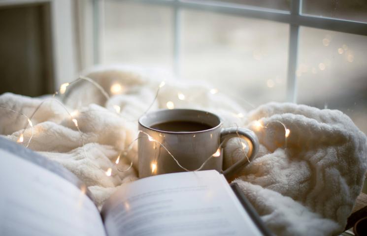 Photo d'une tasse posée sur une couverture et entouré d'une guirlande lumineuse devant une fenêtre avec un livre ouvert au premier plan