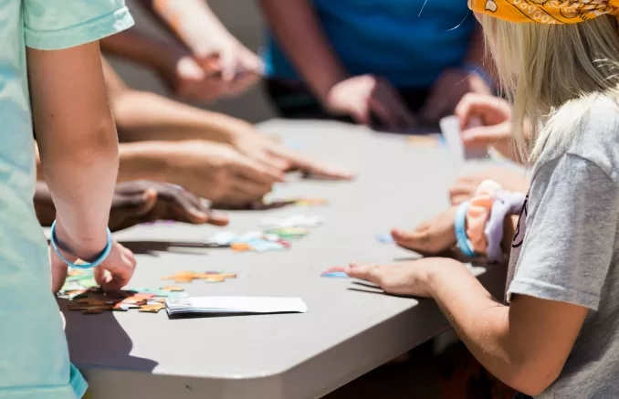 Photo de dos d'un adulte et un enfant qui prennent des pièces de puzzle sur une table. En arrièe plan légèrement flouté, on voit les mains de 5 autres personnes qui sont aussi autour de la table et participent au puzzle.